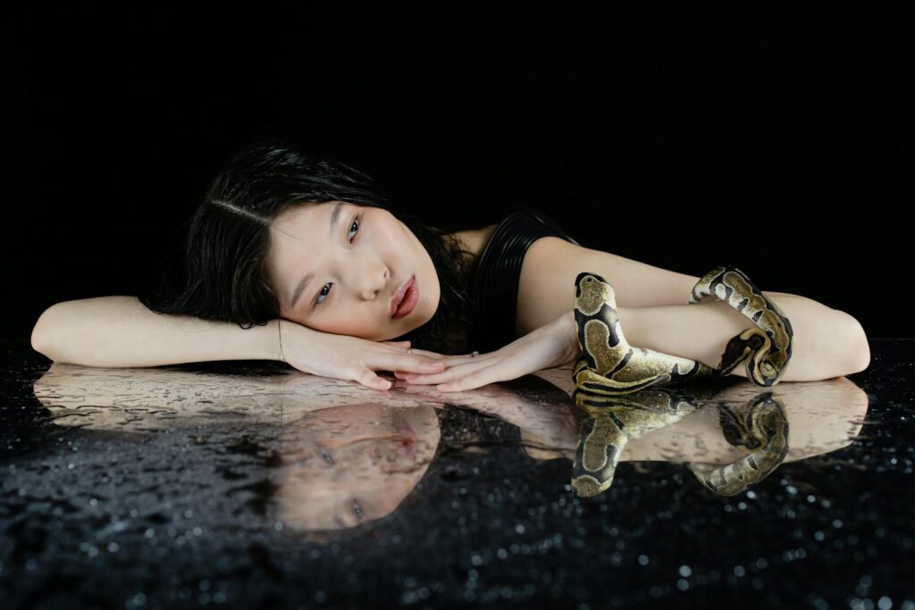 A woman in a dark setting with a snake on a reflective surface, creating a mystical and eerie vibe.